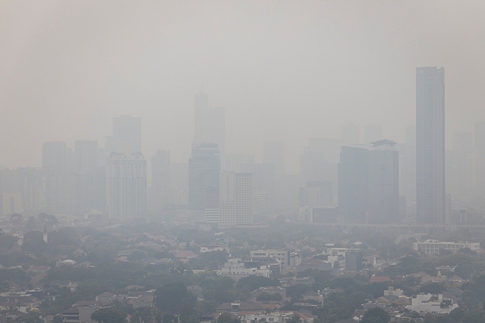 A city skyline obscured by heavy smog. Smaller residential buildings are visible in the foreground, with greenery dotted about. High-rise buildings stand at the back, barely visible through the cloud.